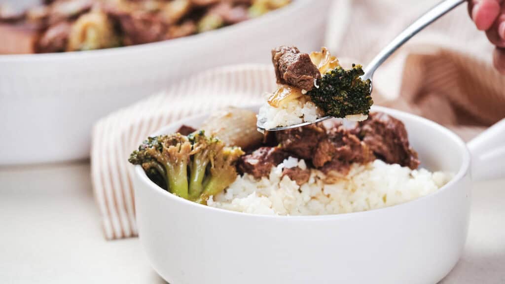 A person serving a bowl of beef and broccoli over white rice with a spoon.