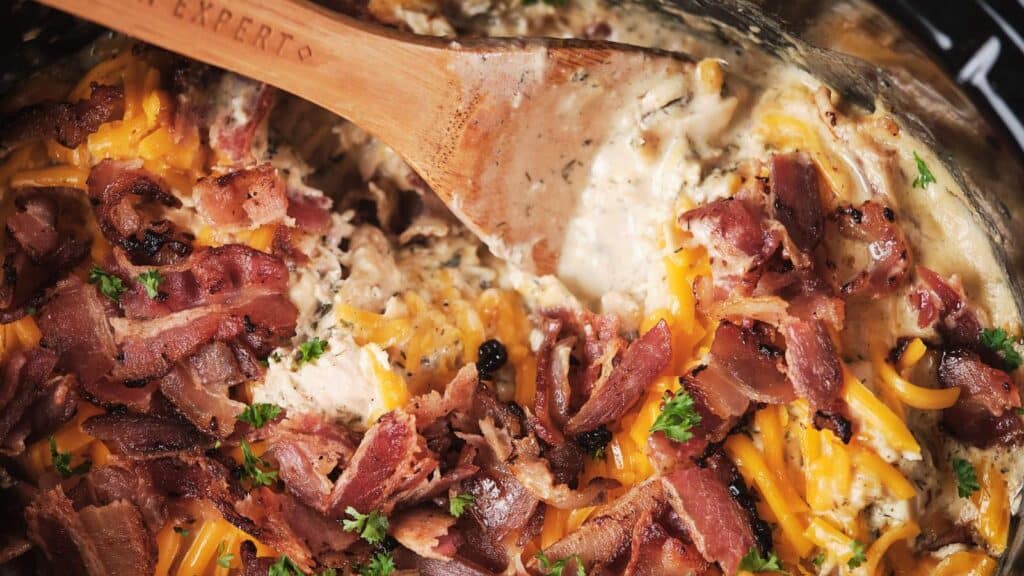 A skillet containing a creamy, cheesy chicken dish garnished with crispy bacon pieces and parsley, stirred with a wooden spoon., Ceamy, cheesy chicken dish garnished with crispy bacon pieces and parsley, stirred with a wooden spoon.