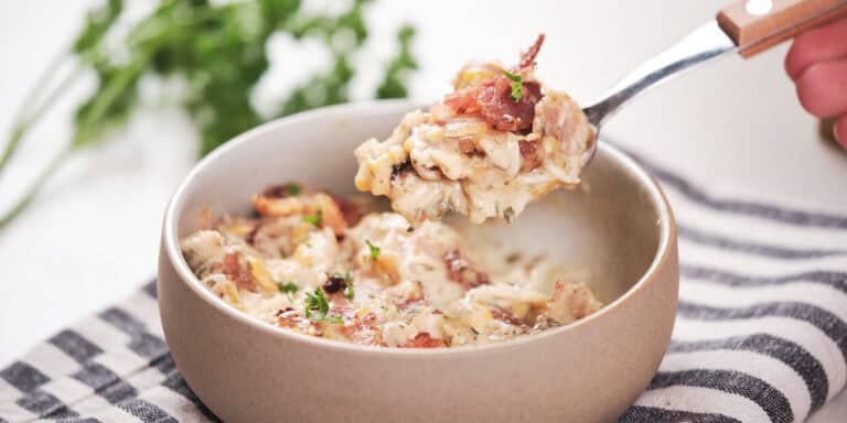A fork lifts a serving of creamy chicken topped with bacon and garnished with herbs from a ceramic bowl.