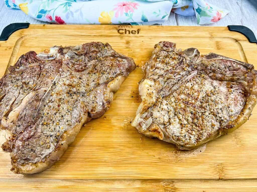 Two cooked T-bone steaks seasoned with spices are placed on a wooden cutting board. A floral cloth is partially visible in the background.