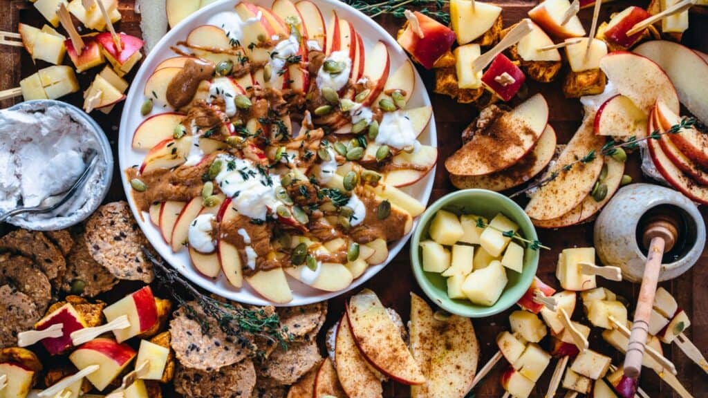 A platter of apple slices topped with sauces and seeds is surrounded by various cheeses, crackers, and apple cubes on skewers.