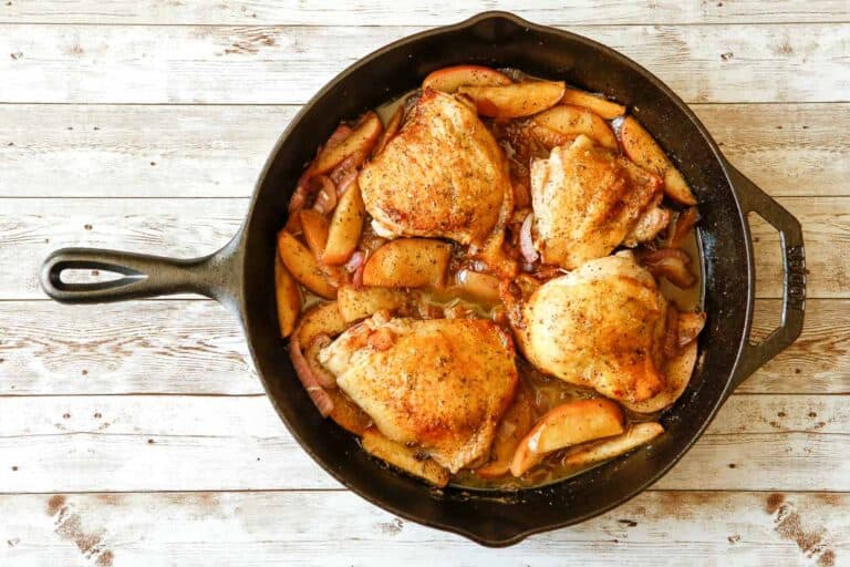 Overhead view of a cast iron skillet containing cooked, seasoned chicken thighs and sliced apples, offering a delicious meal with less mess. The skillet is placed on a wooden surface.