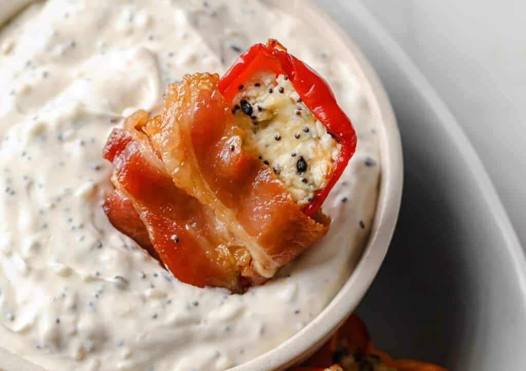 Close-up of a small red pepper stuffed with white creamy filling and black seeds, wrapped in bacon, placed atop a bowl of creamy dip with visible black seeds.