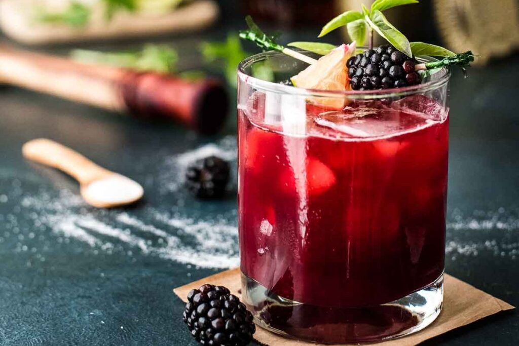 A glass of dark red, quick and delicious cocktail garnished with blackberries and herbs sits on a table with a decanter, golden shaker, and small bowl of lime slices in the background.