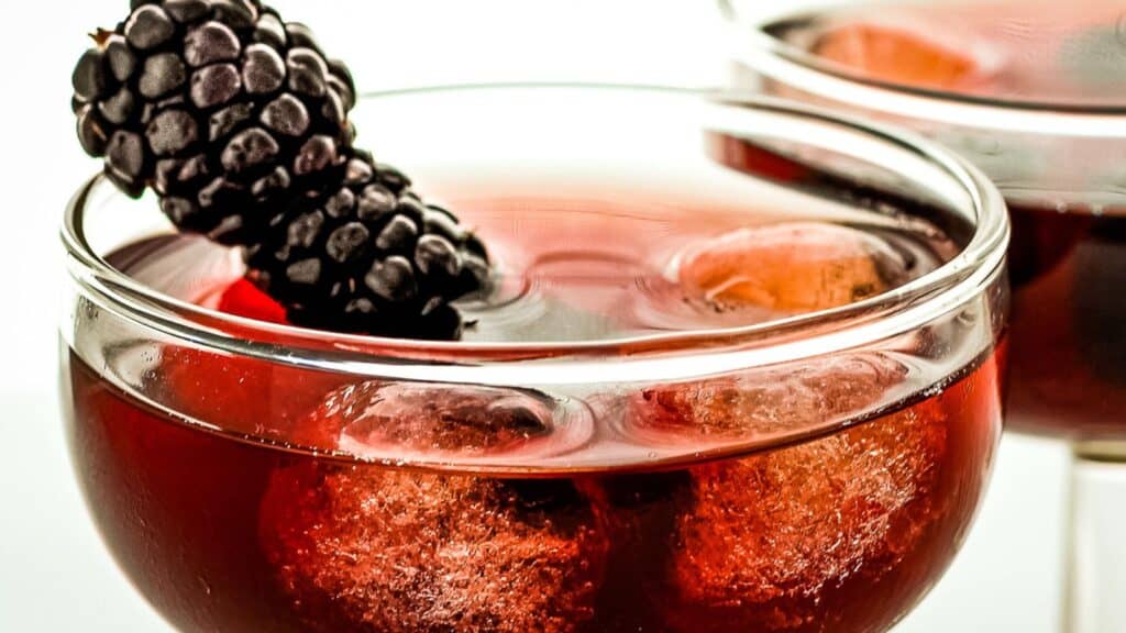 A close-up of a glass filled with a red beverage, ice cubes, and garnished with a blackberry. Another similar glass is partially visible in the background.