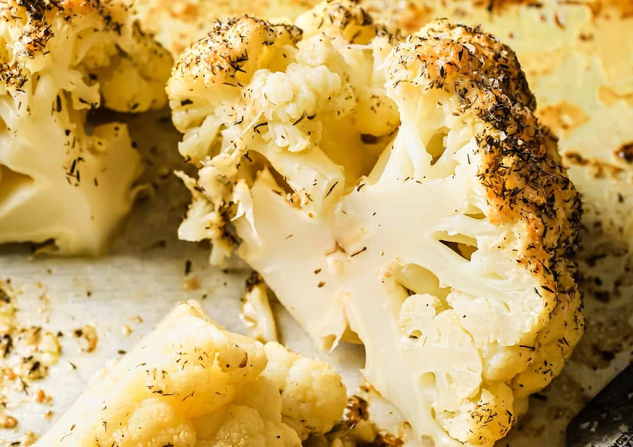 A roasted whole cauliflower head topped with herbs and seasoning sliced in half on a baking tray.
