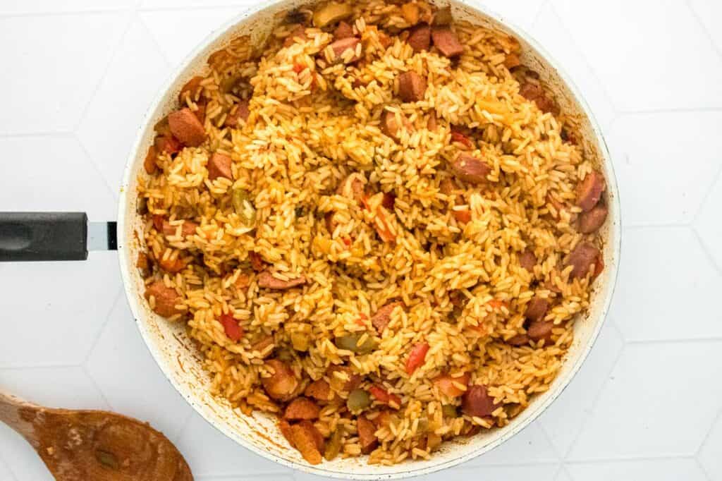 A pan filled with cooked rice mixed with vegetables and small chunks of meat, offering a convenient, less mess meal. A wooden spoon rests beside the pan on a white surface.