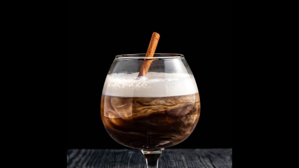 A glass of iced coffee with a layer of foam on top, stirred with a cinnamon stick, set against a black background.