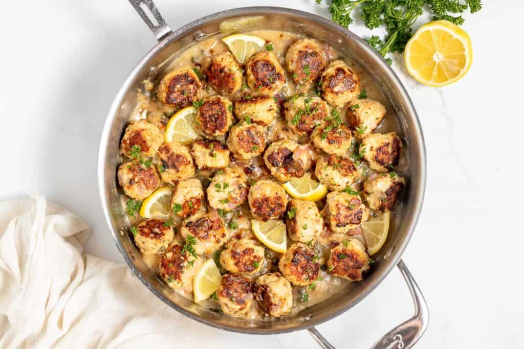 A pan of cooked meatballs in a creamy sauce, garnished with chopped parsley and lemon wedges.