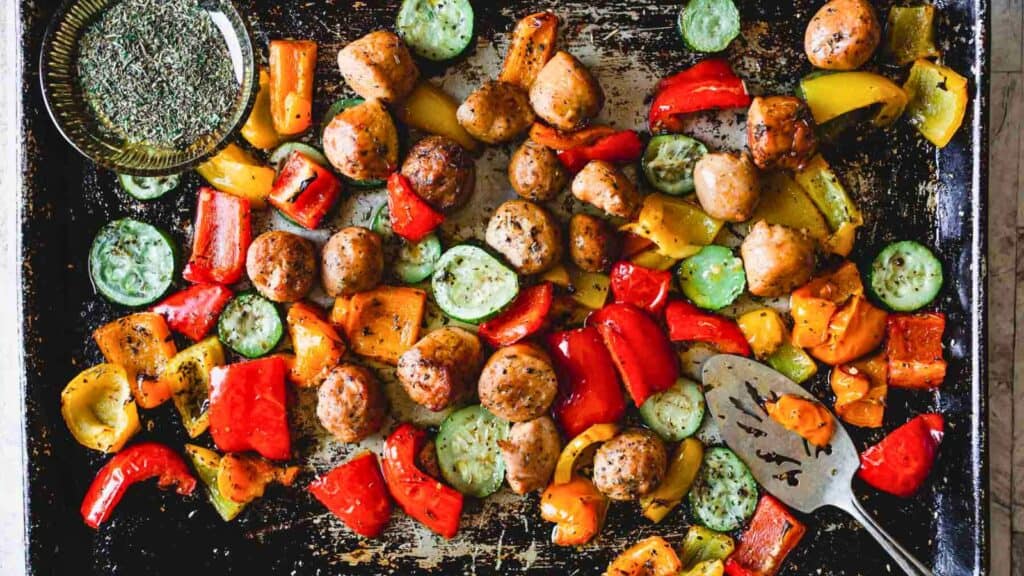 A baking sheet with roasted sausage, sliced zucchini, and red, yellow, and orange bell peppers. A spatula and a small bowl of herbs are also visible.