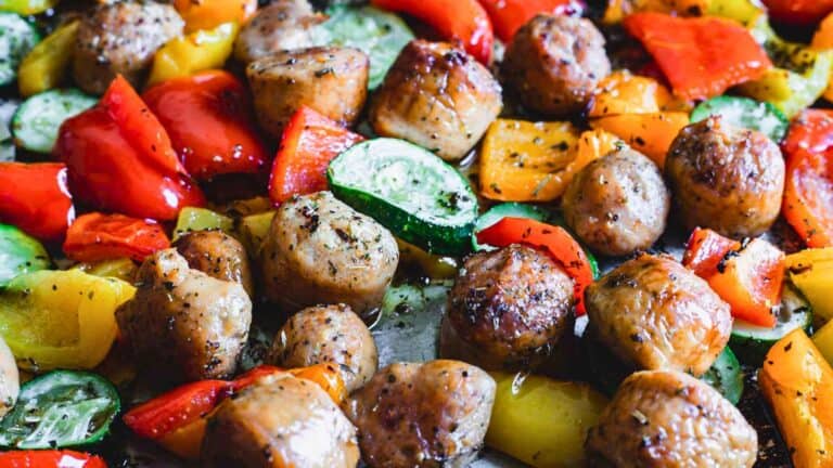 Close-up of cooked sausage pieces with red and yellow bell peppers and sliced zucchini on a tray.