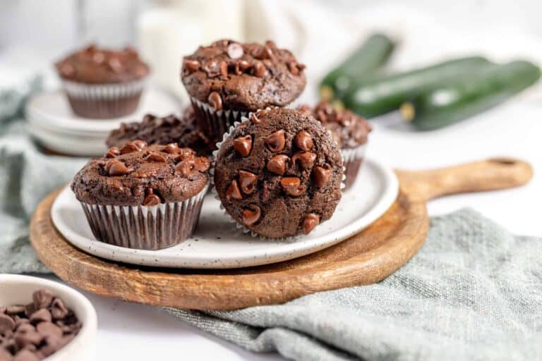 A plate of chocolate muffins and quick breads topped with chocolate chips is displayed on a wooden board, with some zucchinis and a bowl of extra chocolate chips in the background.