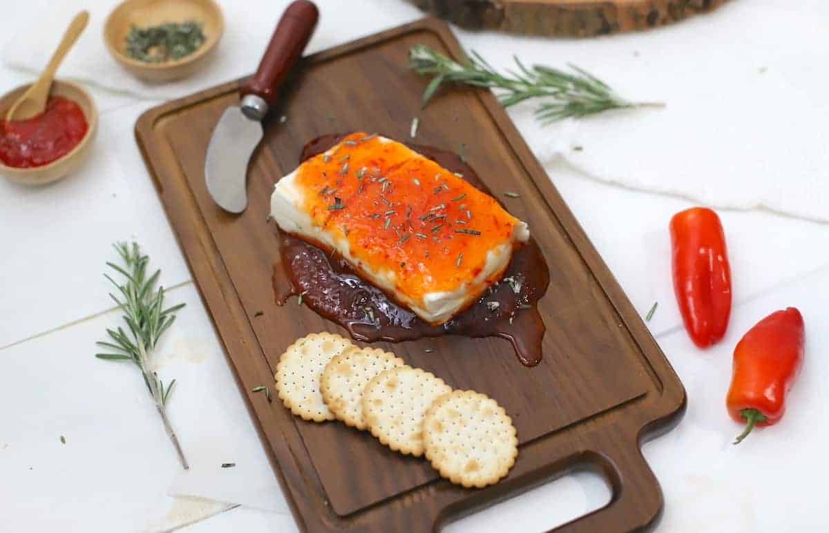 A wooden board with cream cheese topped with a red sauce, garnished with rosemary, and accompanied by round crackers. A knife, herbs, and red peppers are placed beside it.