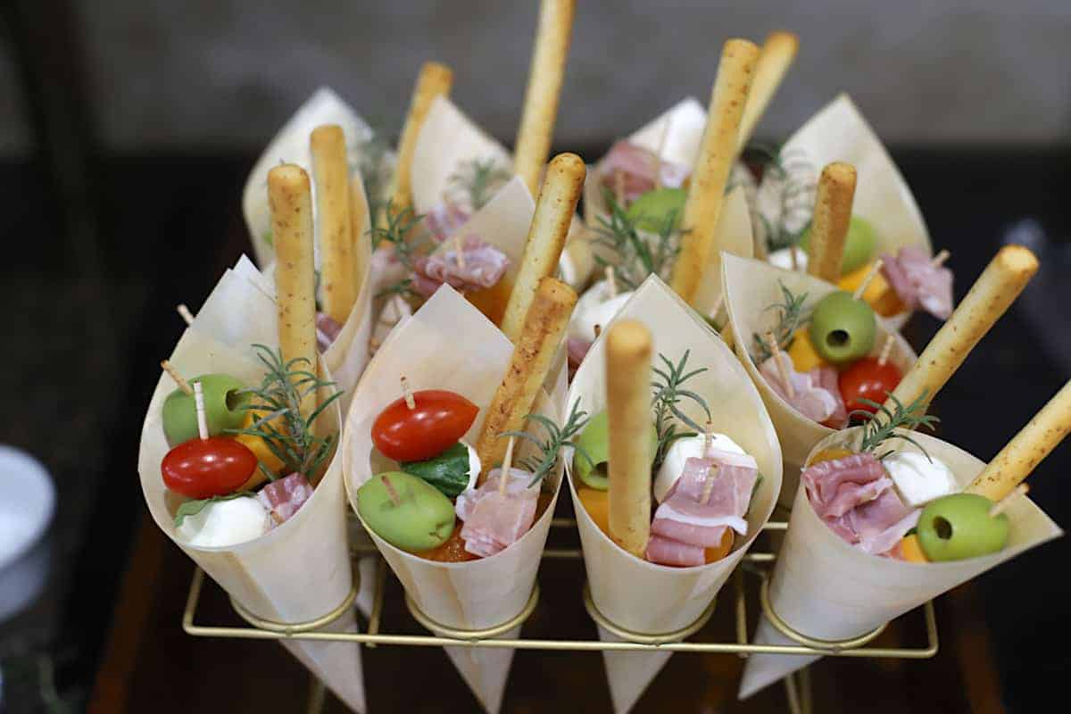Cone-shaped appetizers featuring breadsticks, cherry tomatoes, green olives, cheese, and meat slices, garnished with sprigs of rosemary, arranged on a gold rack.