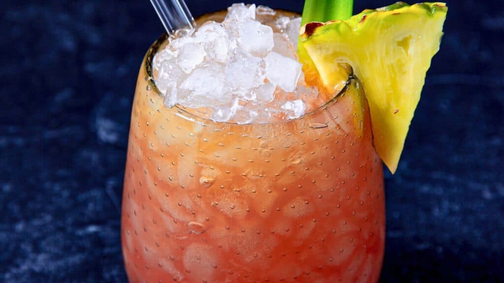 A tropical cocktail garnished with a pineapple slice and a straw, filled with ice, shown against a dark background.