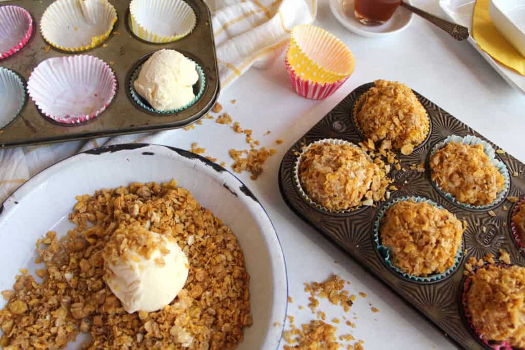 Two baking trays hold empty cupcake liners and cornflake-coated ice cream. A bowl contains crumbled cornflakes and a scoop of vanilla ice cream. Scattered cornflakes and a napkin are on the table.