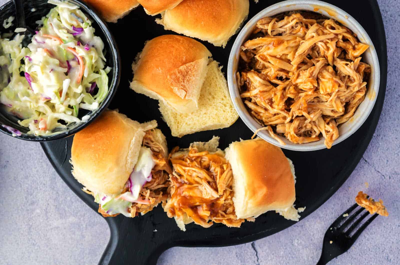A wooden plate holds six sliders filled with shredded chicken and coleslaw, arranged neatly on a surface.