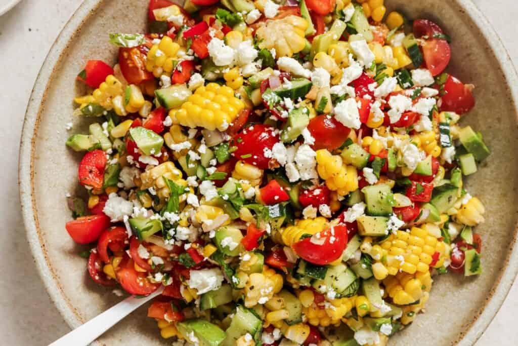 A bowl of colorful salad featuring corn, diced cucumber, cherry tomatoes, chopped avocado, and crumbled feta cheese, garnished with fresh herbs.