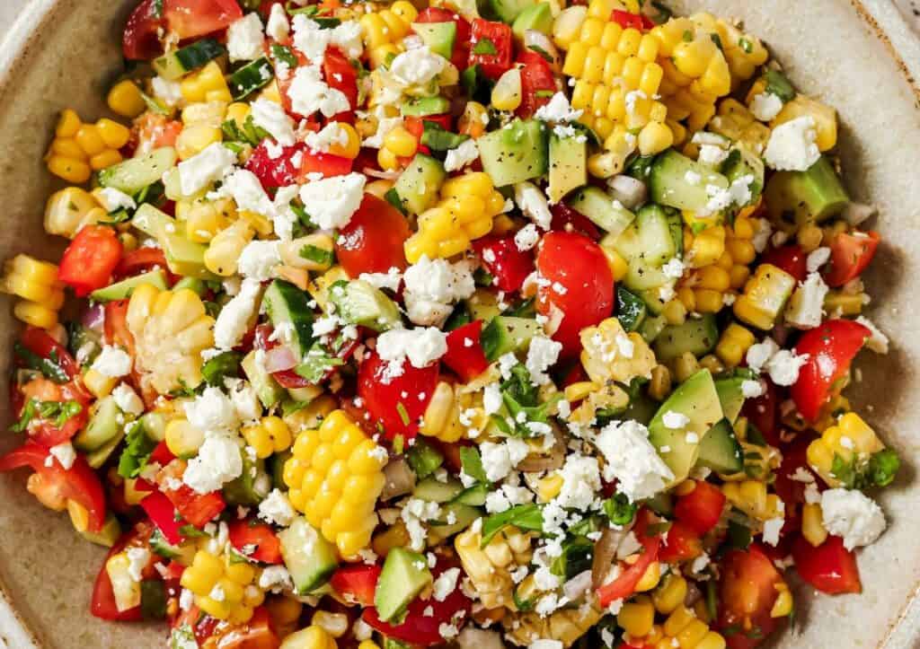 A close-up of a colorful salad featuring corn, cherry tomatoes, cucumber, bell pepper, avocado, and crumbled feta cheese, all served in a brown bowl.