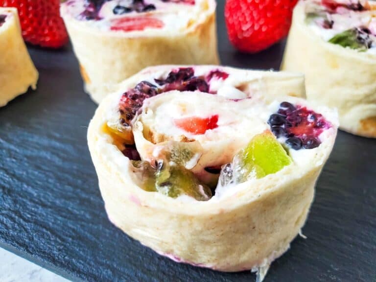 Close-up of a fruit wrap roll, featuring visible pieces of kiwi, strawberries, and blackberries, arranged on a dark slate surface.