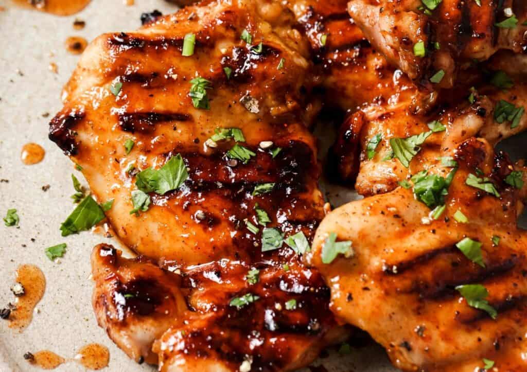 Grilled chicken thighs garnished with chopped herbs and black pepper, shown on a beige plate, with visible grill marks and sauce.