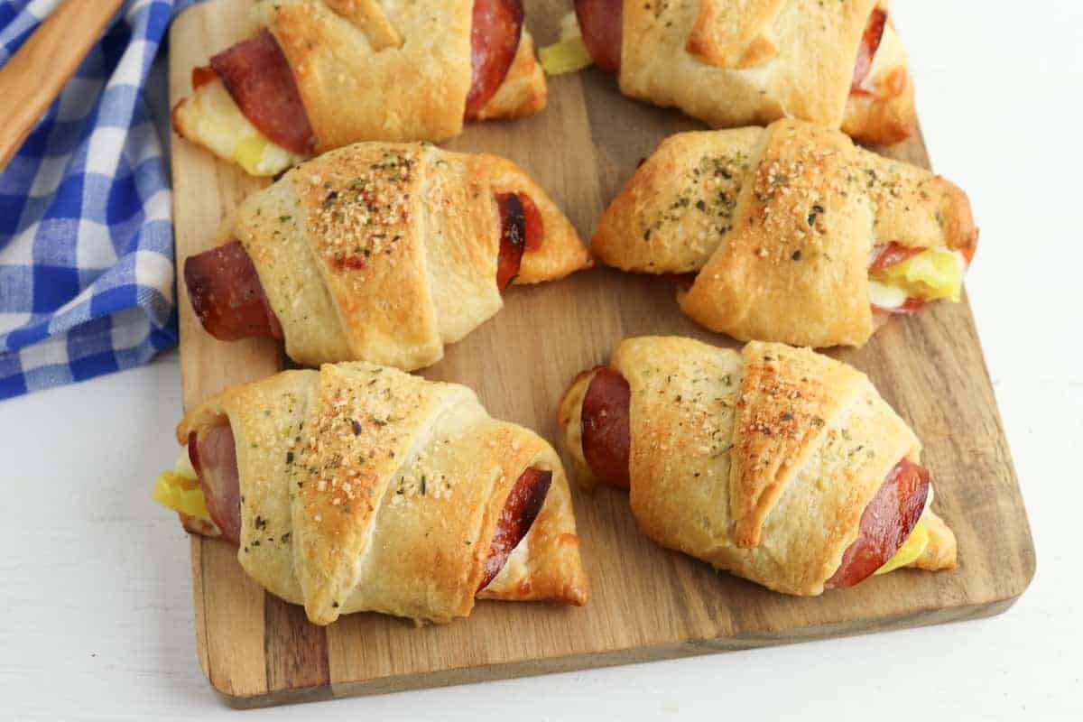 Six baked pepperoni and cheese crescent rolls on a wooden cutting board next to a blue and white checkered cloth.