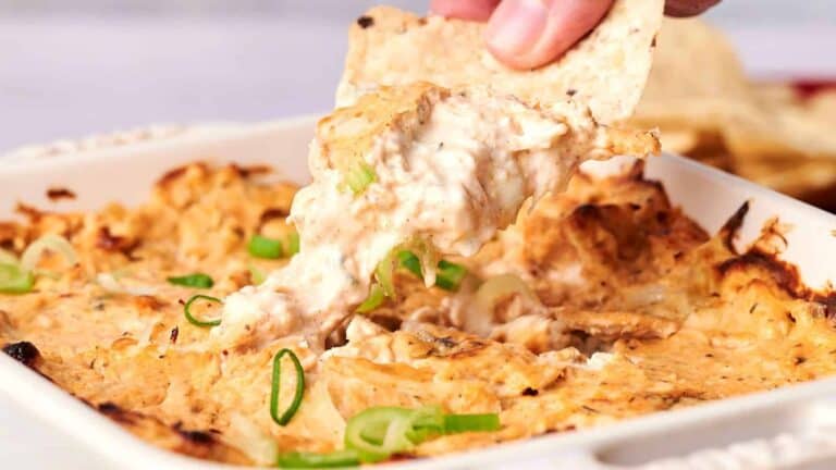 Close-up of a hand dipping a chip into a baked, creamy, cheese and chicken dip garnished with sliced green onions in a white casserole dish.