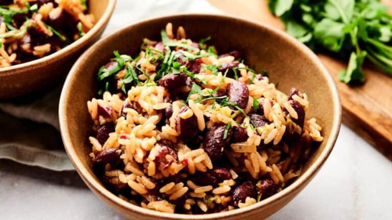 A bowl of rice mixed with red beans and garnished with chopped herbs. Another bowl and a cutting board with fresh green herbs are visible in the background.