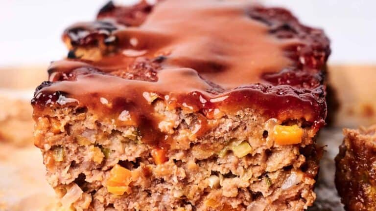 Close-up of a sliced meatloaf with a glossy sauce coating. The meatloaf contains visible pieces of vegetables.