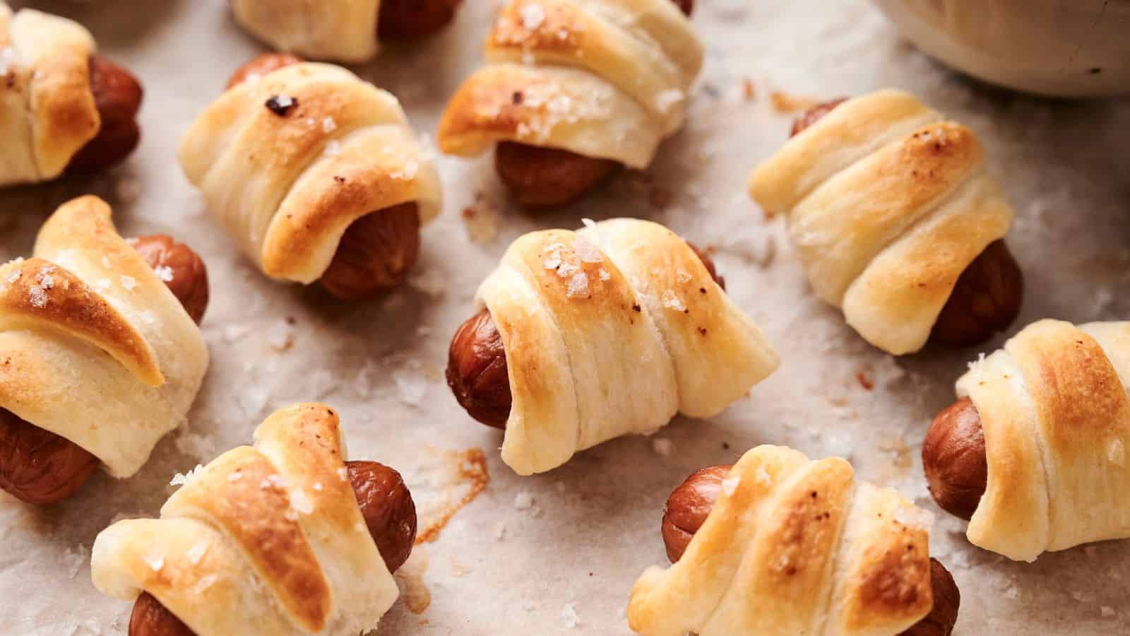 Pigs in a Blanket, small sausages wrapped in puff pastry, baked until golden brown, and sprinkled with coarse salt are displayed on a baking sheet lined with parchment paper.