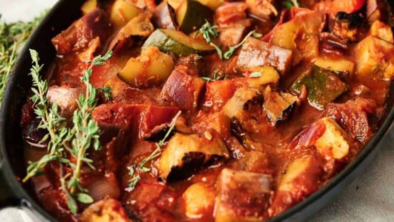 A close-up of a baked vegetable dish with zucchini, eggplant, tomatoes, and fresh thyme, served in a black skillet.