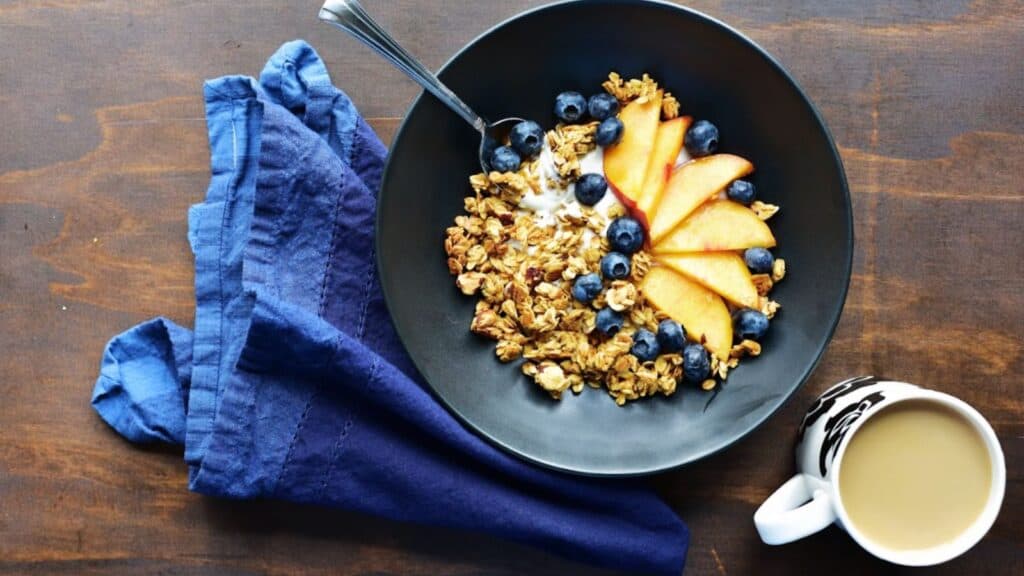 A bowl of granola with blueberries, sliced peaches, and yogurt, accompanied by a cup of coffee, and a folded blue napkin on a wooden table.