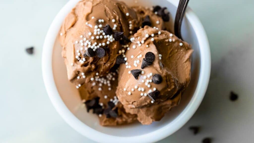 A bowl of chocolate ice cream with chocolate chips and white sprinkles, with a spoon on the side.