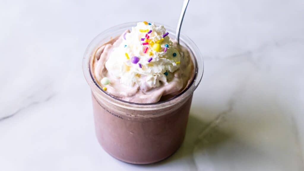 A cup of chocolate milkshake topped with whipped cream, colorful sprinkles, and a spoon on a marble surface.