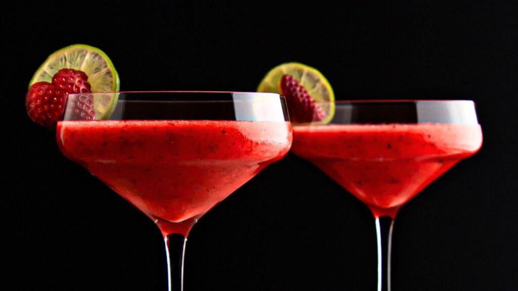 Two red frozen cocktails in martini glasses, garnished with lime slices and strawberries, set against a black background.