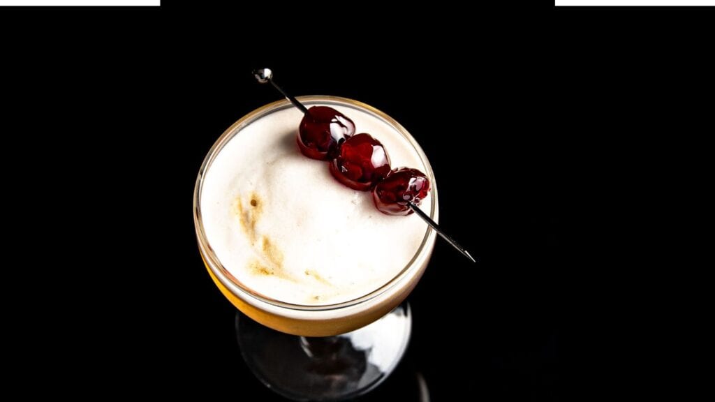 A frothy cocktail garnished with three cherries on a skewered pick, set against a black background.