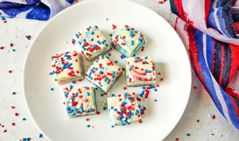 A white plate contains six pieces of white chocolate fudge decorated with red, white, and blue sprinkles. The plate is surrounded by red, white, and blue fabric and scattered sprinkles.