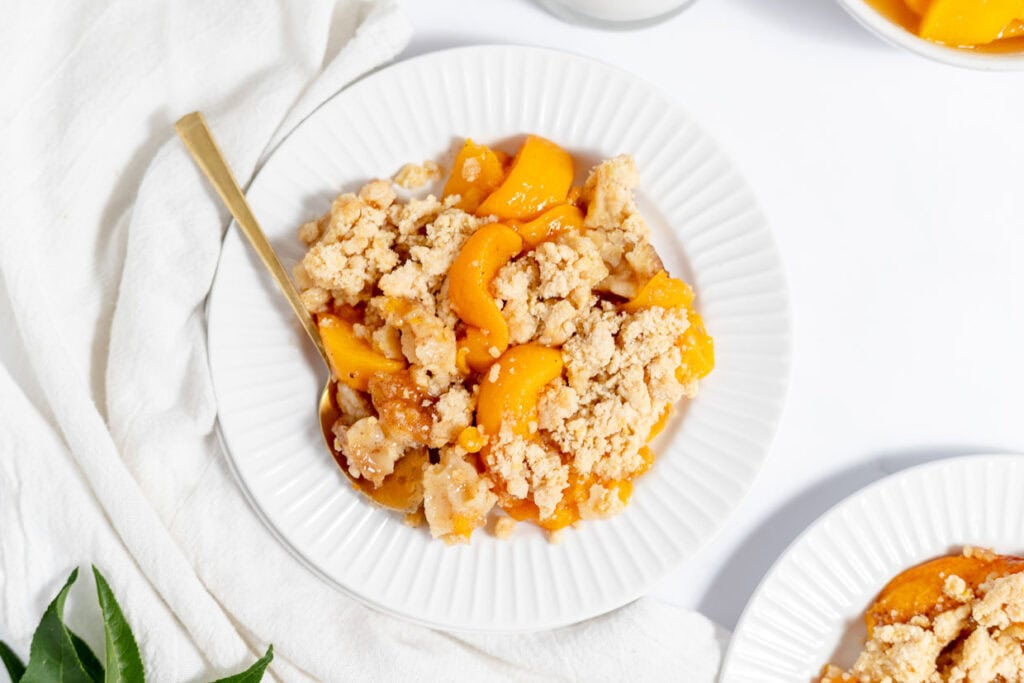 A white plate holds a serving of peach cobbler with a golden-brown crumb topping and a fork resting on the side. The background includes a white cloth and a hint of greenery.