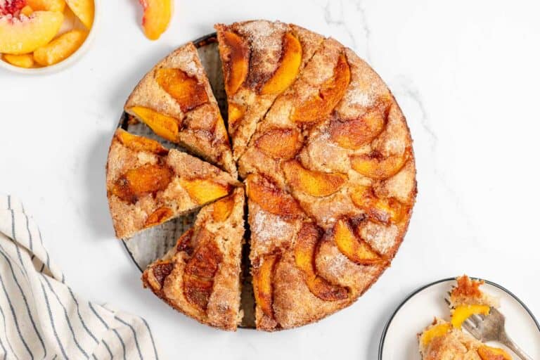 A round peach cake sliced into eight pieces on a white surface. A few slices of peach are in a small bowl on the upper left, and a fork with a piece of cake is on a plate on the lower right. The setup is simply peachy keen, with every detail inviting you to indulge.