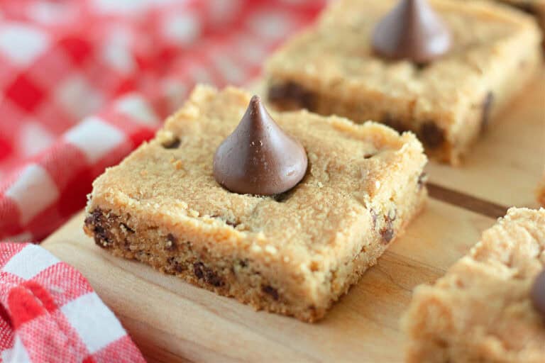 Close-up of a cookie bar topped with a chocolate kiss, set on a wooden surface with a checkered red and white cloth in the background.