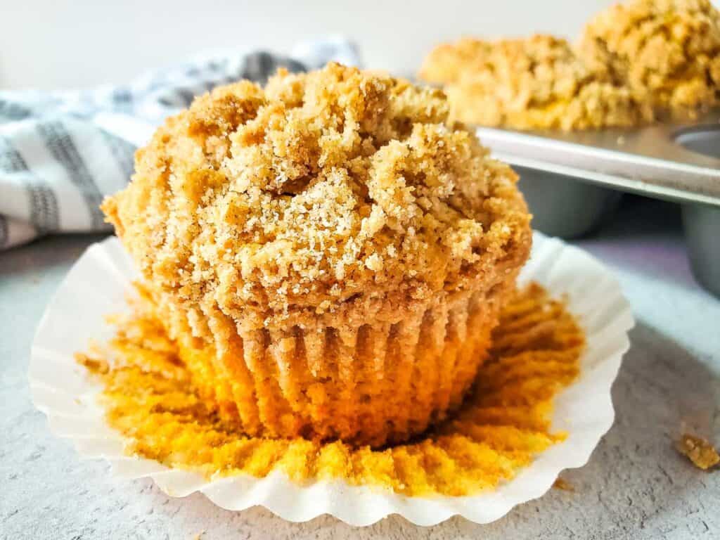 A muffin with a crumb topping partially unwrapped from its paper liner, with more muffins in a baking tray in the background.