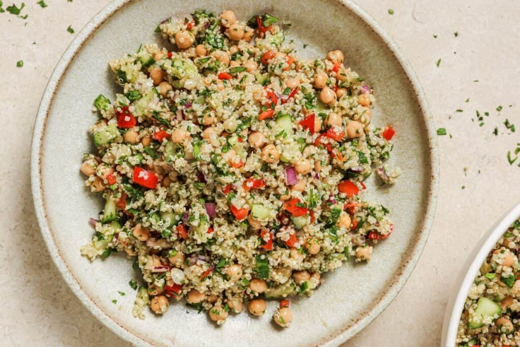 A bowl filled with a quinoa salad featuring chickpeas, chopped red bell peppers, cucumbers, red onions, and fresh parsley.