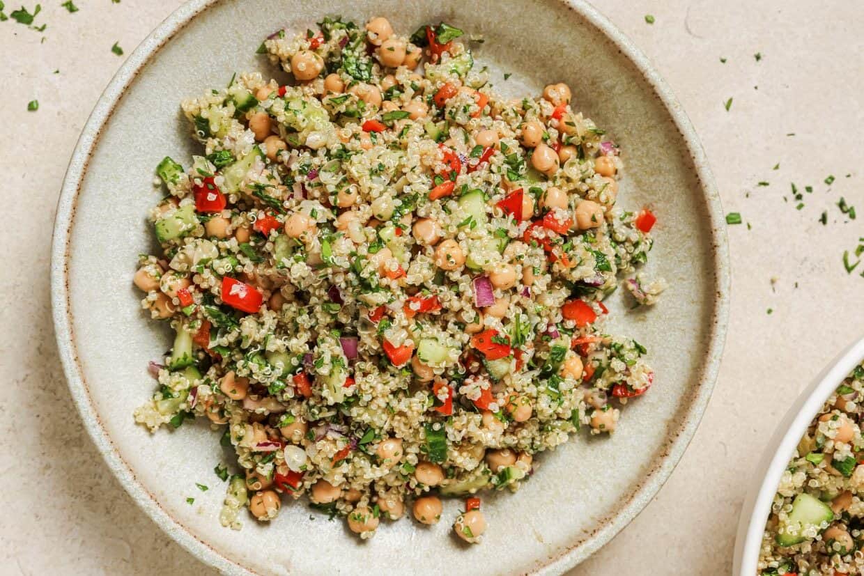 A bowl filled with a quinoa salad that includes chickpeas, chopped red bell peppers, cucumbers, red onions, and fresh herbs, presented on a light-colored surface.