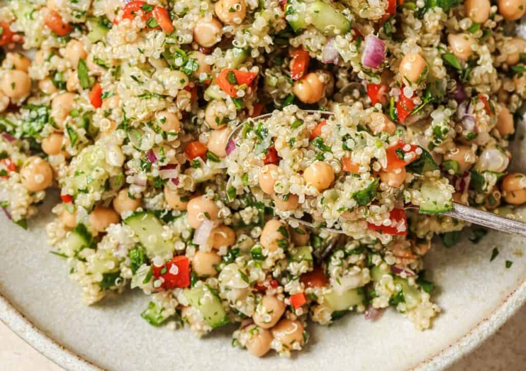 Close-up of a quinoa salad with chickpeas, cucumbers, red peppers, red onions, and parsley. A spoon is scooping a portion from the dish.