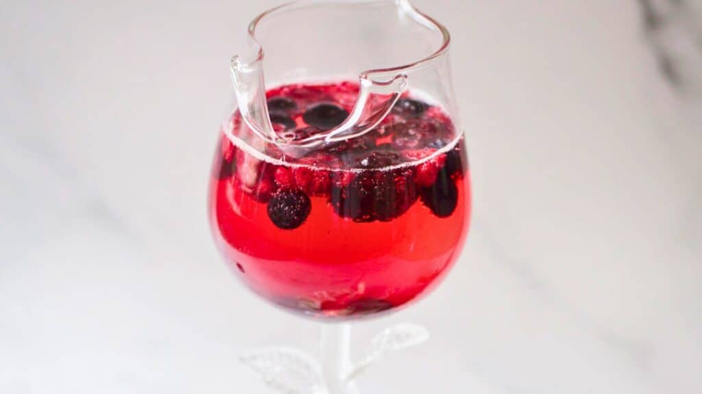 A partially broken wine glass filled with a red berry-infused drink on a light gray background.