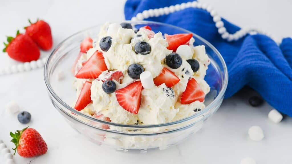 A glass bowl filled with a dessert made of whipped cream, strawberries, blueberries, and mini marshmallows. Several strawberries and blueberries are scattered on the table nearby.