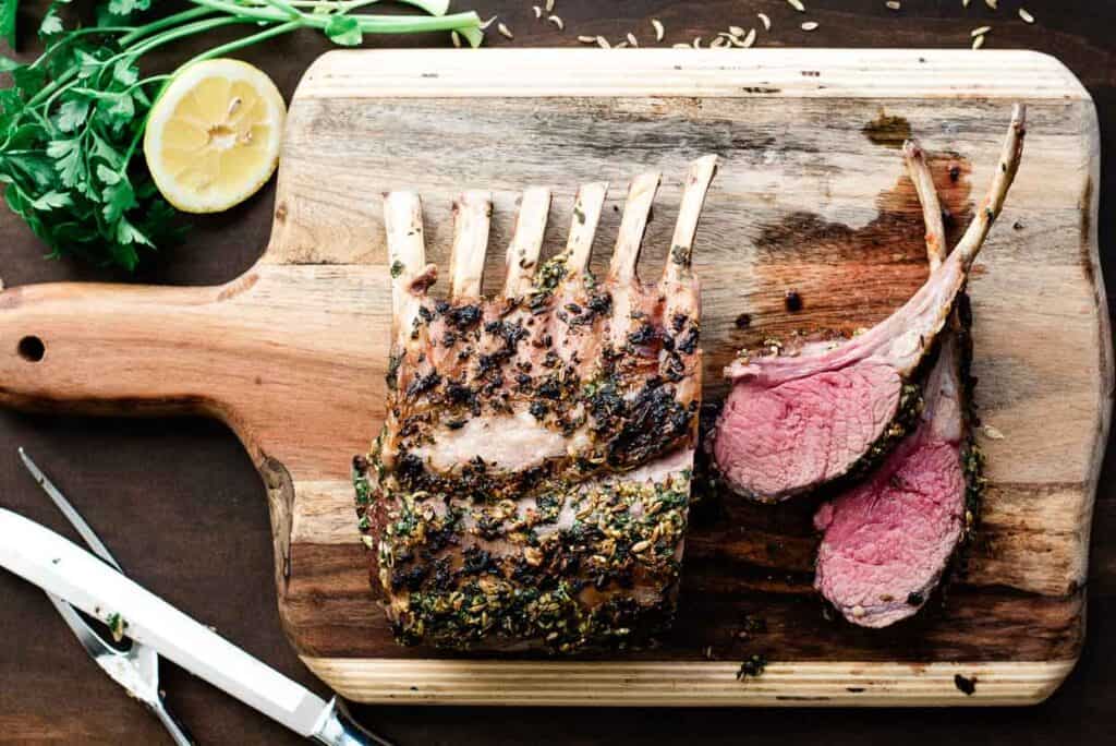 A cutting board holds a cooked rack of lamb seasoned with herbs, partially sliced into two pieces. Lemons and greens are on the side.