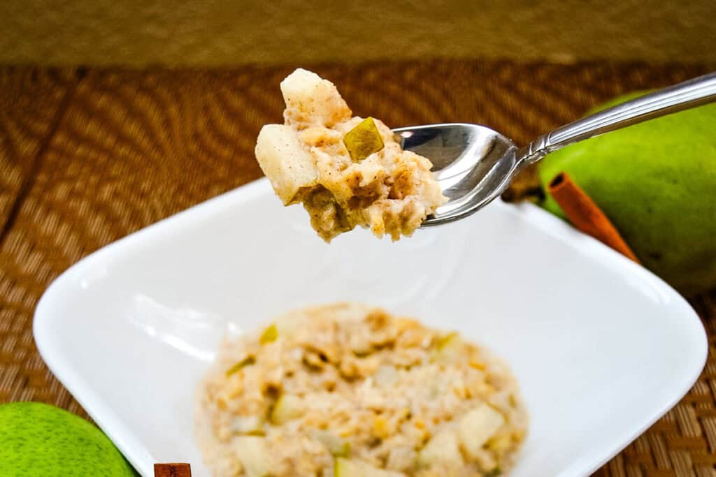 A spoonful of oatmeal with diced apples is held over a white bowl of oatmeal, with a green pear and a cinnamon stick in the background.