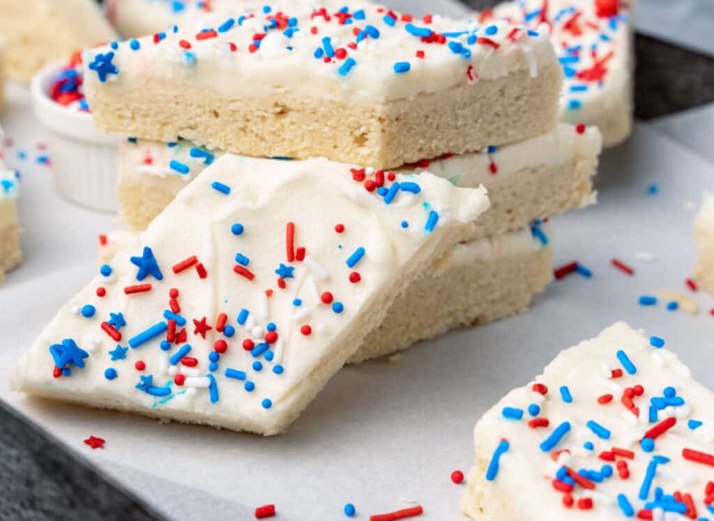 Frosted sugar cookie bars topped with red, white, and blue sprinkles are stacked on a piece of parchment paper.