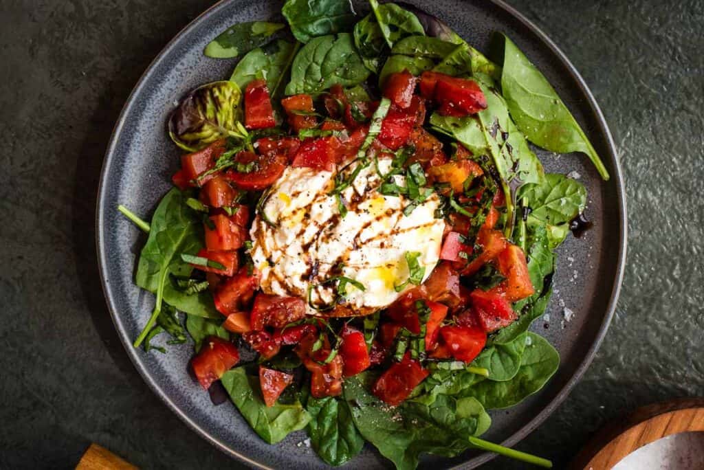 A plate of fresh spinach, diced tomatoes, and a ball of burrata cheese drizzled with balsamic glaze.